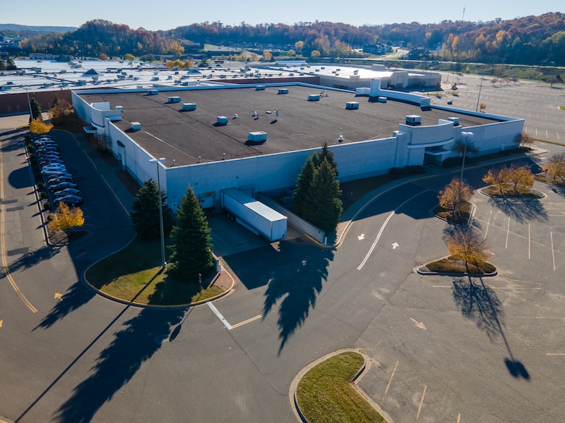 Large Industrial Building With Parking Lot. Semi Trailer Present By Loading Dock. Hvac Present On Roof. Shadows Cast Pine Trees Around Building.  Curves Entry Way.  Colorful Tree Line In Background.