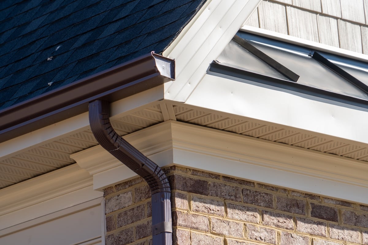 Closeup View Of Dark Brown Gutter System With White Soffit Vent, Gutter Guard, Drop Outlet, Downspout, Vinyl Elbows, Decorative Trim Molding