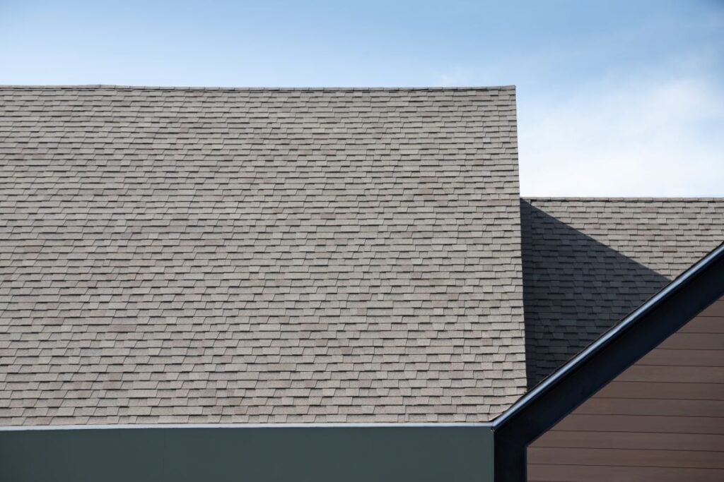 Roof Shingles On Top Of The House Against Blue Sky. Dark Asphalt Tiles On The Roof Background.